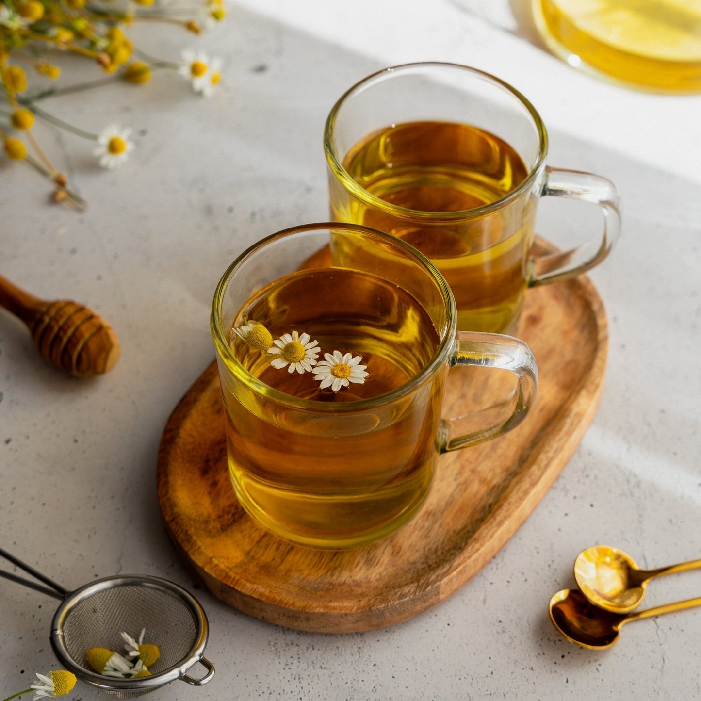 two clear glass mugs of organic chamomile flower tea on wooden tray with chamomile flowers and honey dipper on table