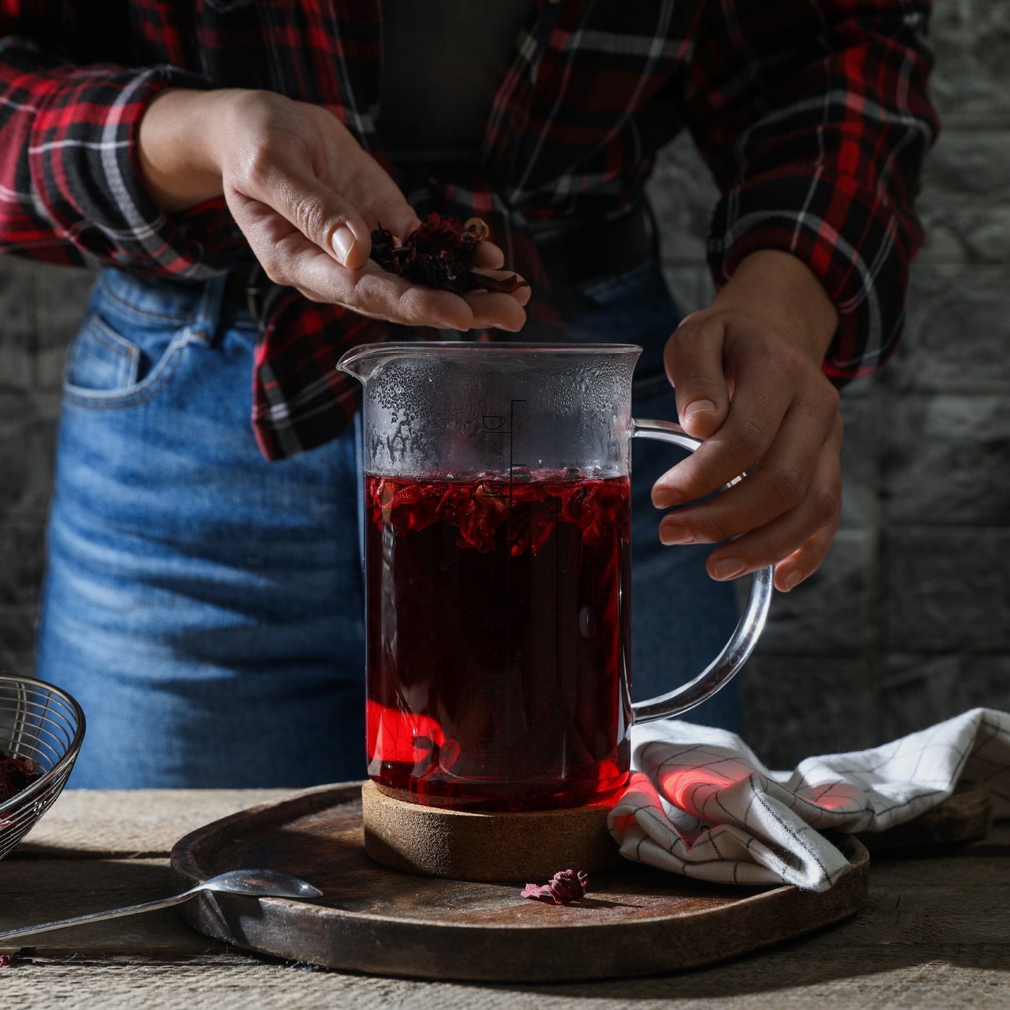Person preparing organic hibiscus flower tea in a clear glass pitcher showing vibrant red infusion